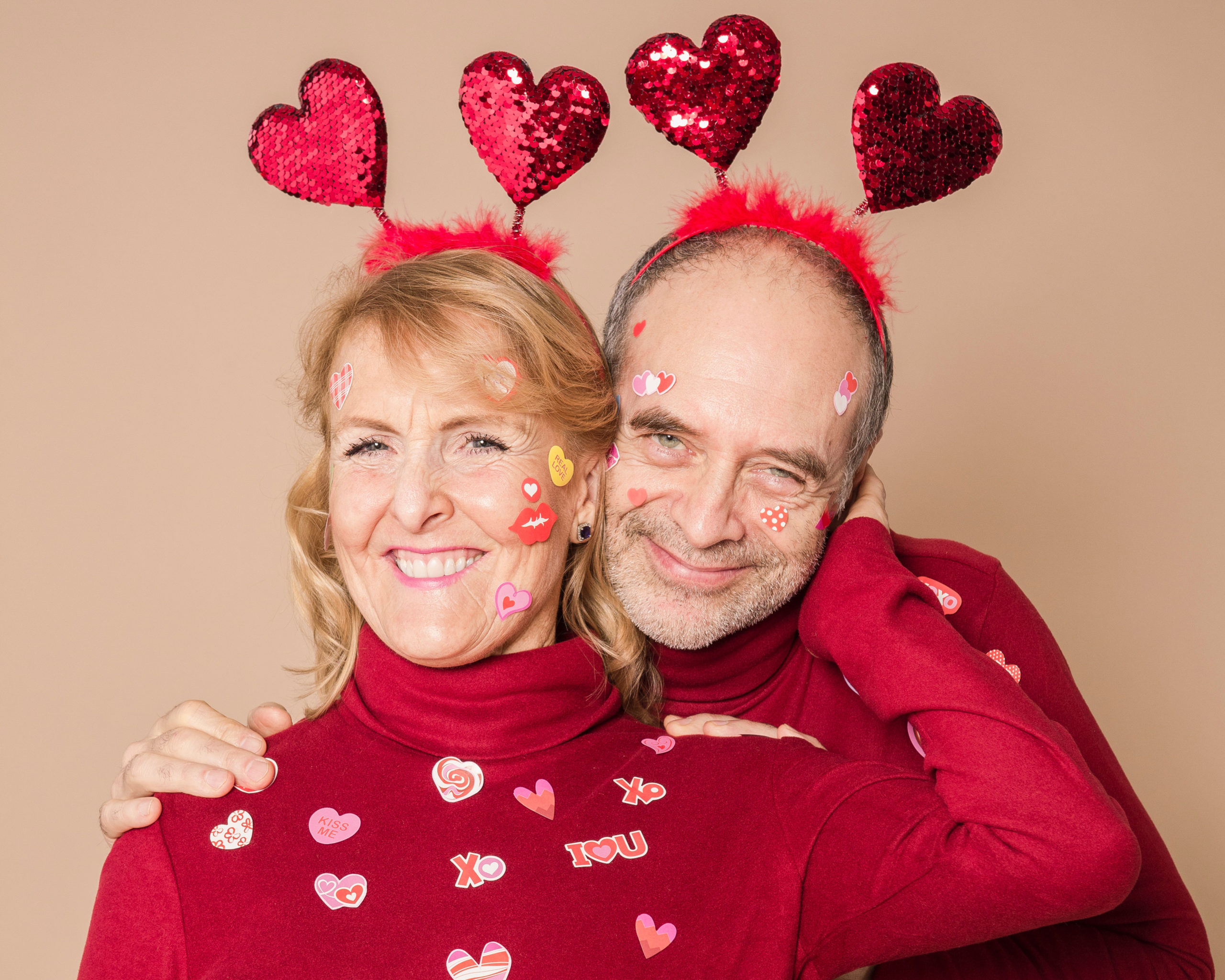 A white couple in their mid-fifties in matching red turtleneck jumpers and love heart bouncy ears. They are cuddling and covered in various 'I love you' stickers.